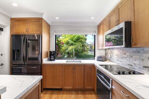 A modern kitchen with wooden cabinets, stainless steel appliances, a large window with a view of greenery, and a tiled backsplash. - Wailea Ekahi 42C - Peekaboo Ocean Views, 2 Full Baths, King Tempurpedic, Full AC (Kihei)