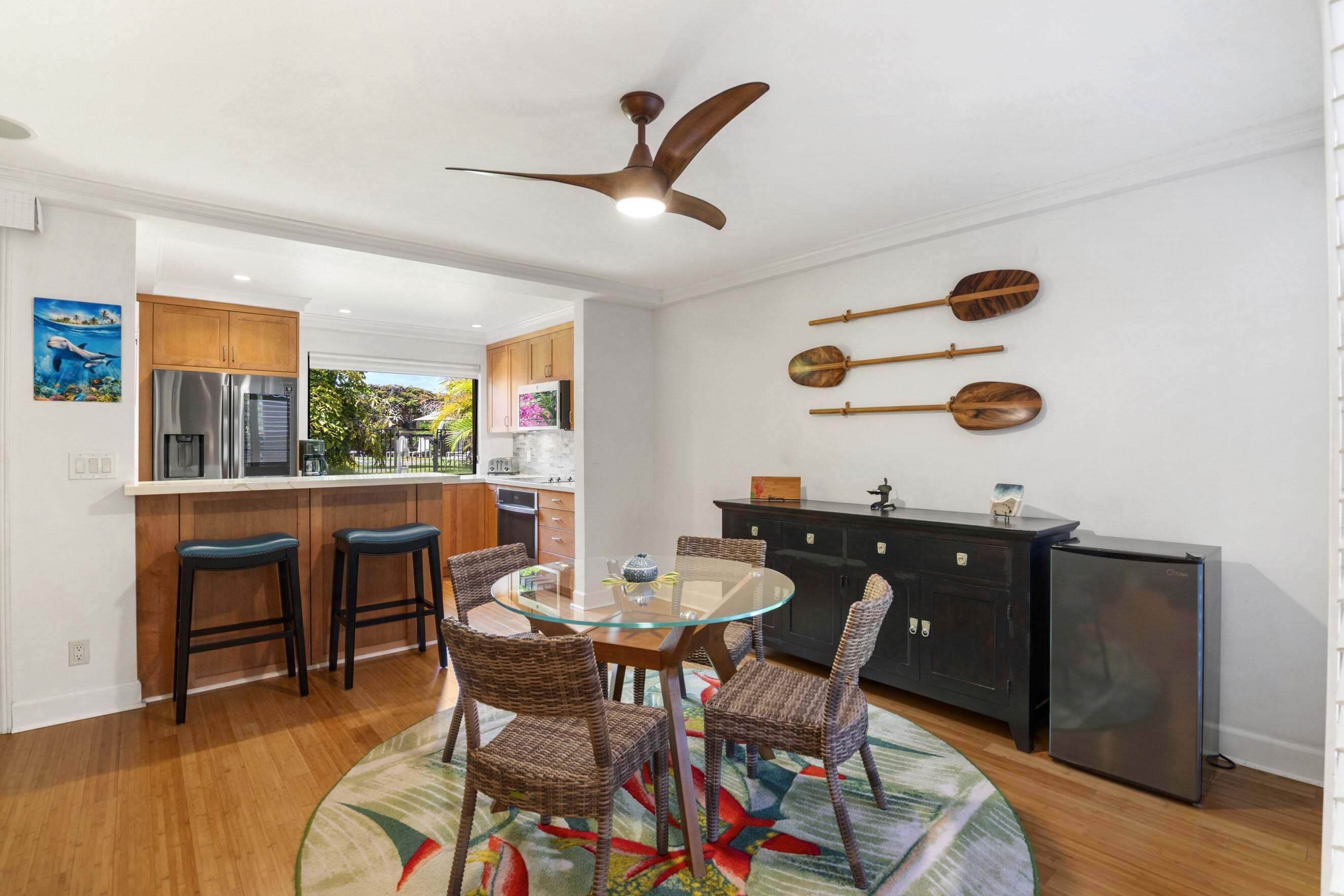A modern kitchen with a glass dining table, wicker chairs, a ceiling fan, and wooden utensils on the wall.