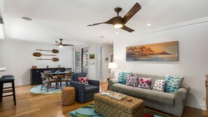 A living room with a sofa, ottoman, dining table, chairs, ceiling fans, and artwork on the wall. - Wailea Ekahi 42C - Peekaboo Ocean Views, 2 Full Baths, King Tempurpedic, Full AC (Kihei)