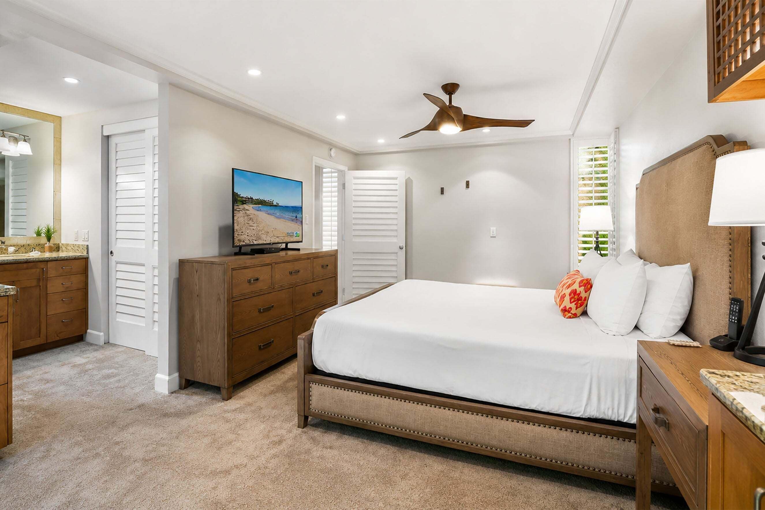 Une chambre à coucher comprenant un lit, une commode, un téléviseur, un ventilateur de plafond et une fenêtre avec des stores.