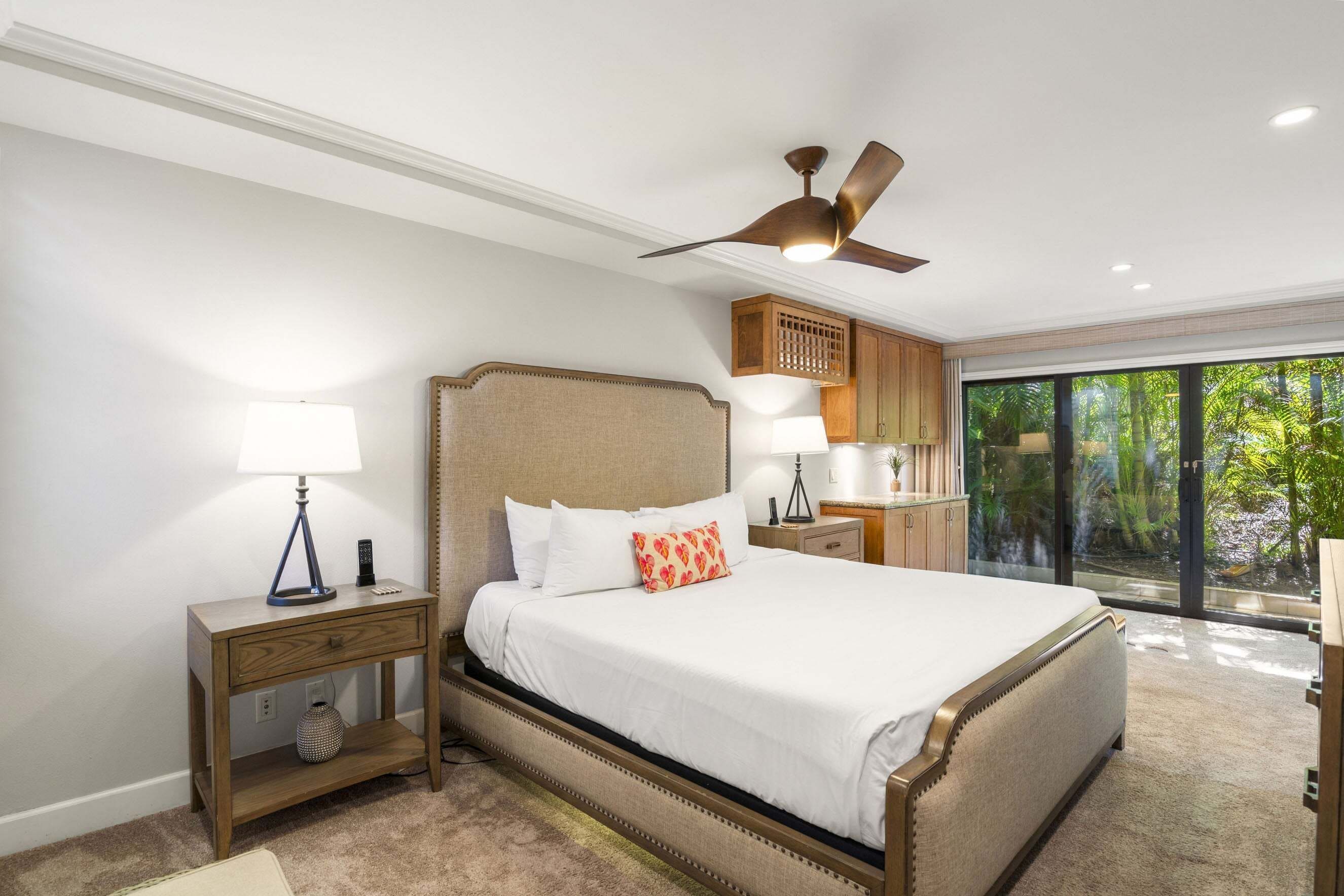 Une chambre à coucher avec un lit, des tables de chevet, un ventilateur de plafond et une vue sur la verdure grâce à de grandes fenêtres.