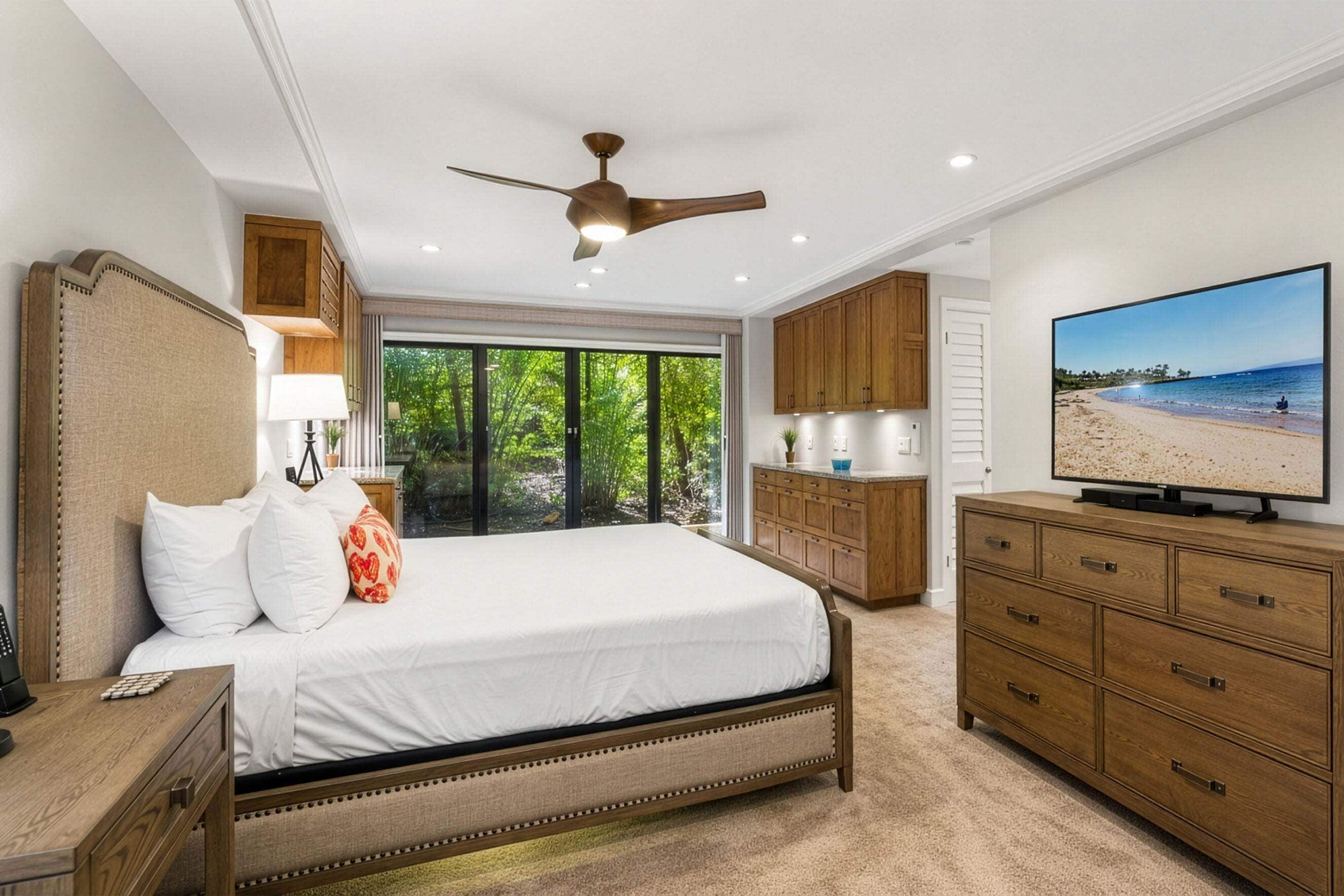 Une chambre à coucher comprenant un lit, un ventilateur de plafond, un téléviseur, des armoires en bois et une vue sur la plage, visible à travers de grandes fenêtres.