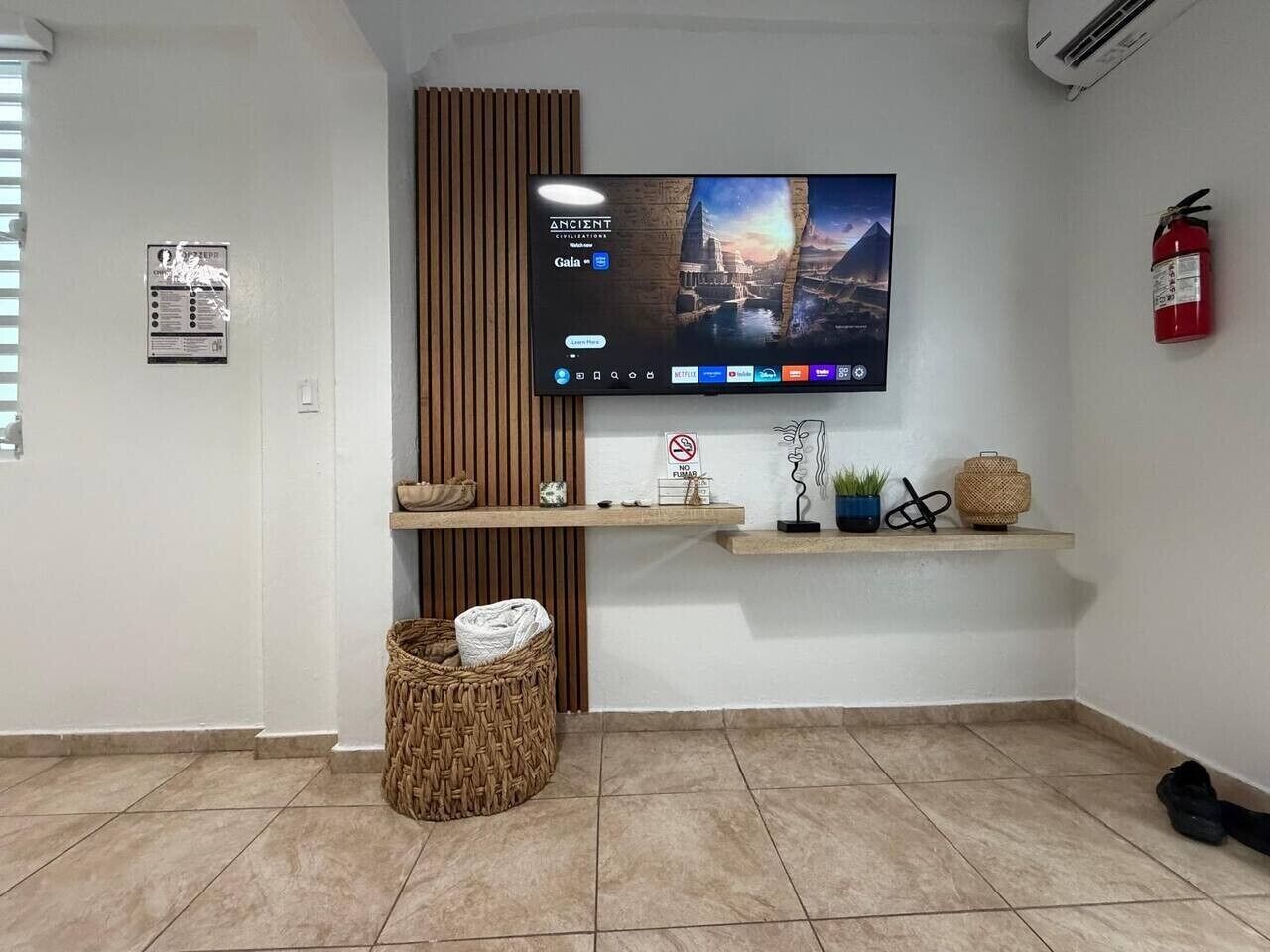 Une pièce avec un téléviseur fixé au mur diffusant un film, un extincteur, un panier en osier et une petite étagère ornée d’objets décoratifs.