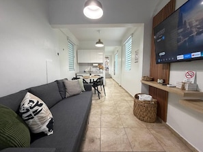 A modern living room with a grey sofa, a flat-screen TV mounted on the wall, and a dining area in the background. - Modern Santurce Apartment Near Beaches for 4 (San Juan de Puerto Rico)