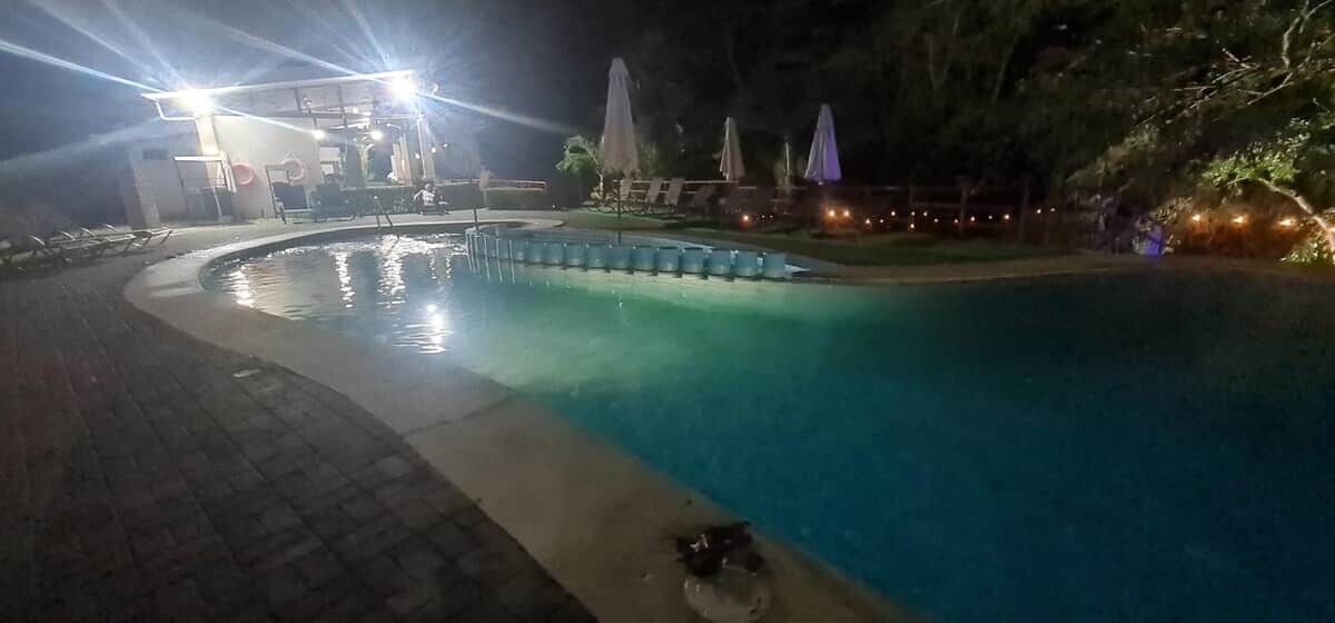 A nighttime swimming pool area with a circular pool, lounge chairs, umbrellas, and a building with lights in the background.