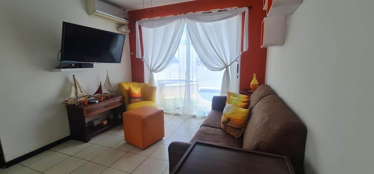 Una sala de estar con televisor de pantalla plana, sofá, sillón, mesita y ventana con cortinas.