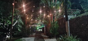 Un chemin bordé d’arbres et illuminé par des guirlandes lumineuses, qui mène à une construction dotée d’un toit.