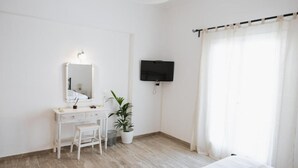A bedroom with a bed, a desk with a chair, a potted plant, a television, and a window with curtains. - White Wave Studios (santorini)
