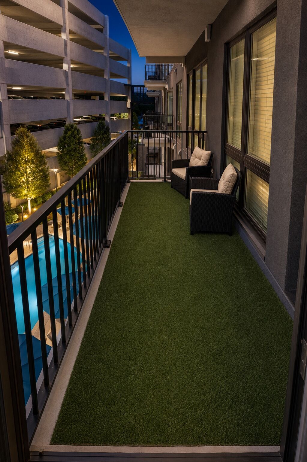 Ein Balkon mit künstlichem Rasen, zwei schwarze Korbstühle mit Kissen, ein Pool und im Hintergrund ein Mehrabsoblagenhaus mit Balkonen.