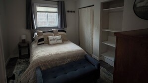 Quarto com cama, ventoinha no teto, janela com cortinas, mesa de cabeceira e guarda-roupa.