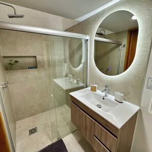 A modern bathroom with a glass-enclosed shower, a vanity with a mirror, and a small shelf with toiletries. - Casa Legaspi, Perfect for Work and Relaxation in GDL (Guadalajara)