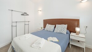Una cama bien hecha con una colcha azul y blanca, dos almohadas estampadas, una mesita de noche blanca con una lámpara y un toallero de pared.