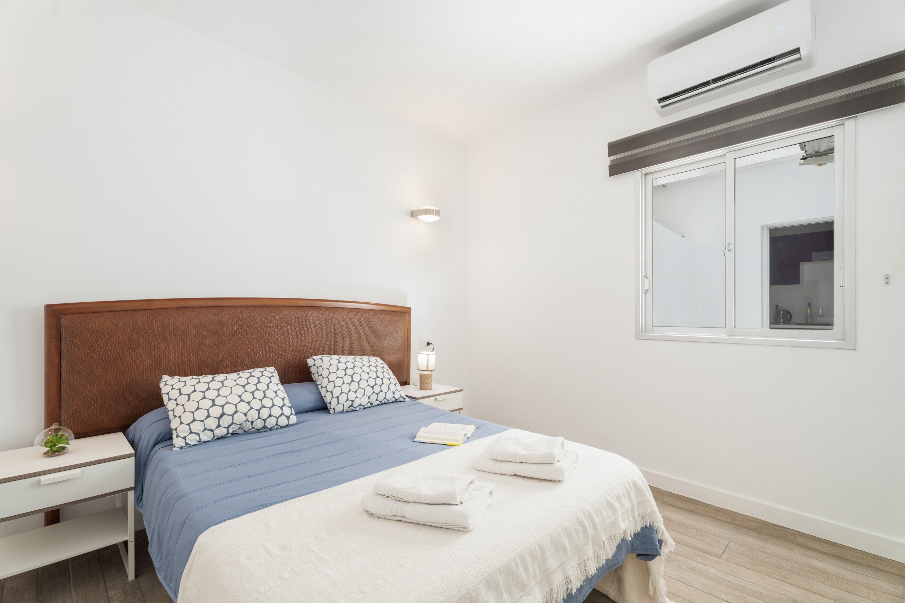 Ein modernes Schlafzimmer mit einem Holzkopfteil, einem Nachttisch mit Lampe, einem Fenster mit Blick und ordentlich gefalteten Handtüchern auf dem Bett.