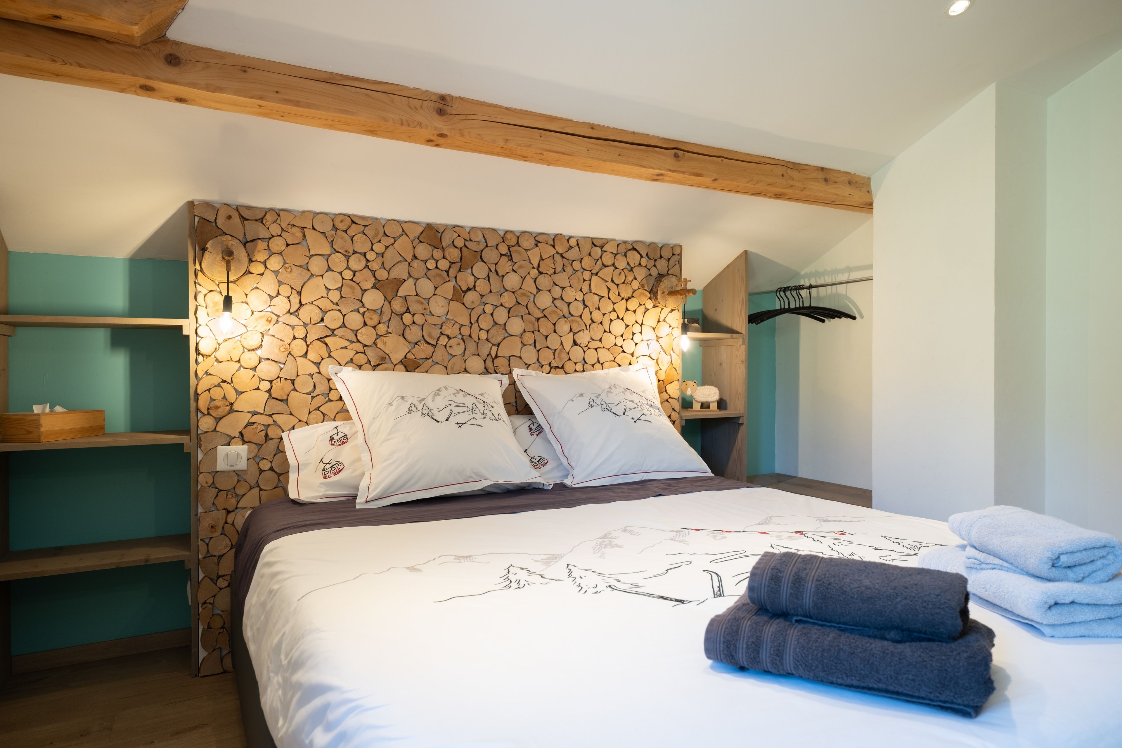 Een slaapkamer met een bed, kussens, een houten hoofdbord, een plank, een kledingroede en een wandlamp.
