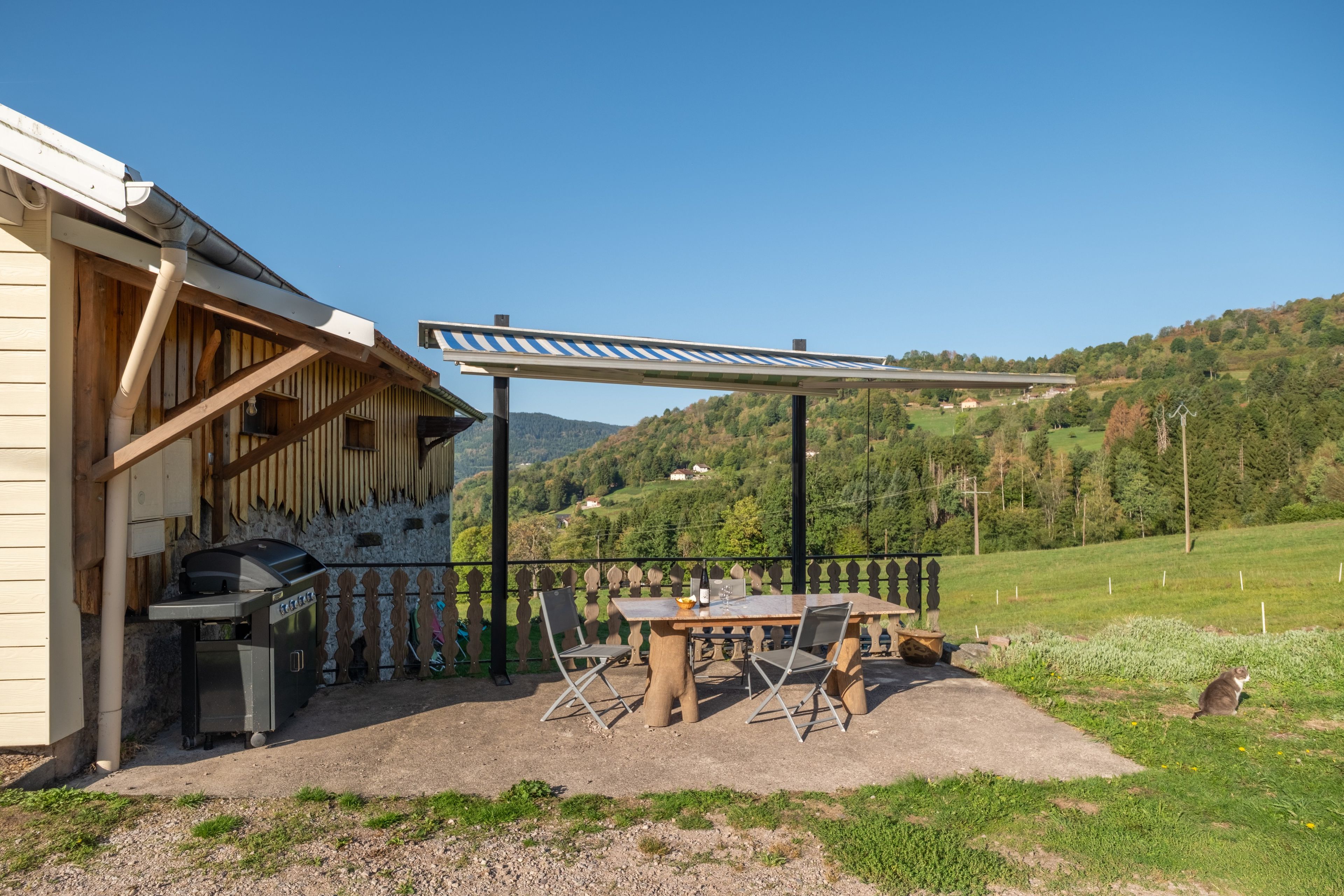 An outdoor patio with a grill, dining table, and chairs, overlooking a hilly landscape.
