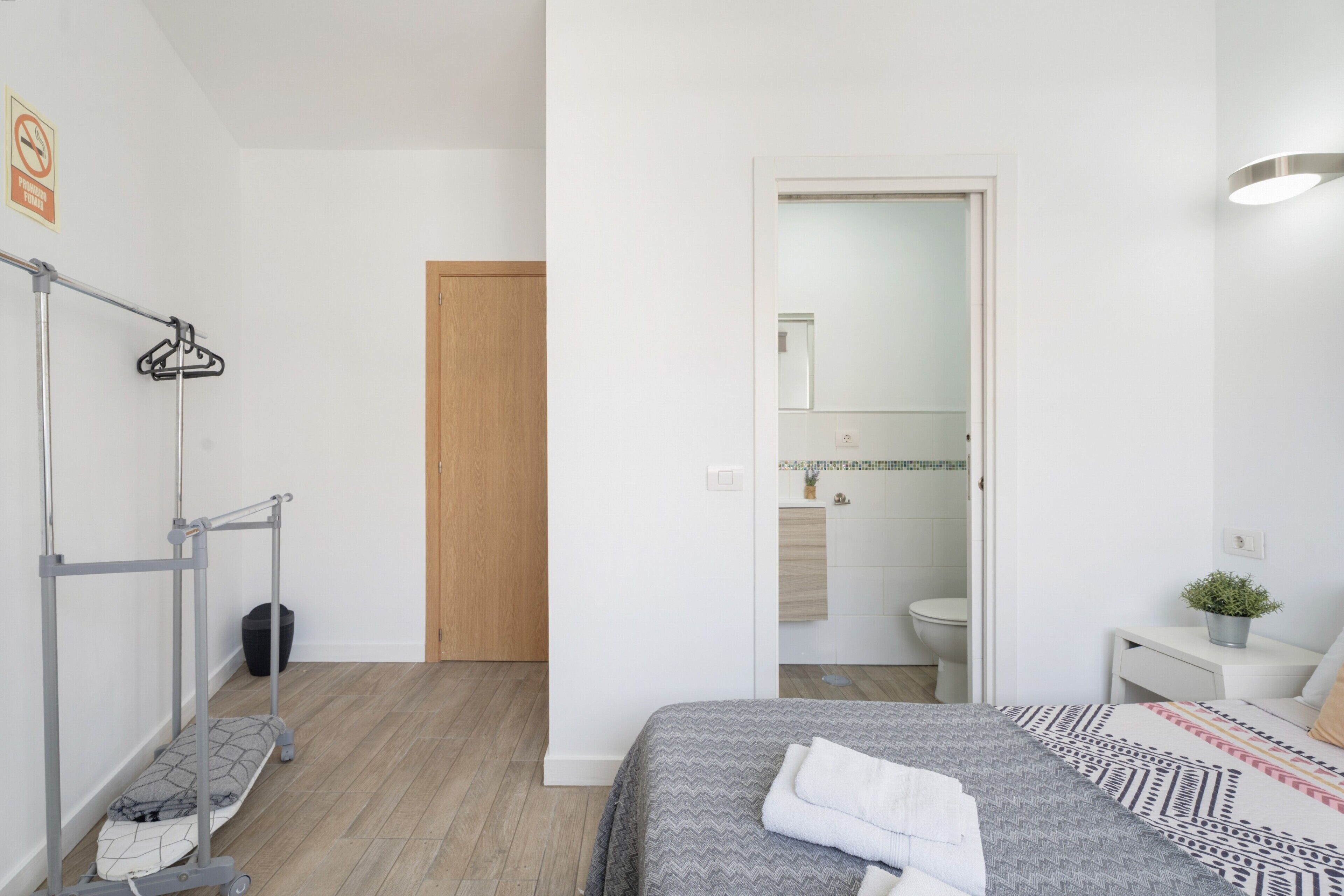 A modern bedroom with a bed, a hanging clothes rack, a wooden door, and a bathroom visible through an open door.