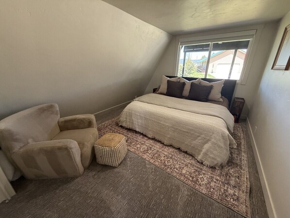 A bedroom with a bed, a chair, a small ottoman, and a window with a view.