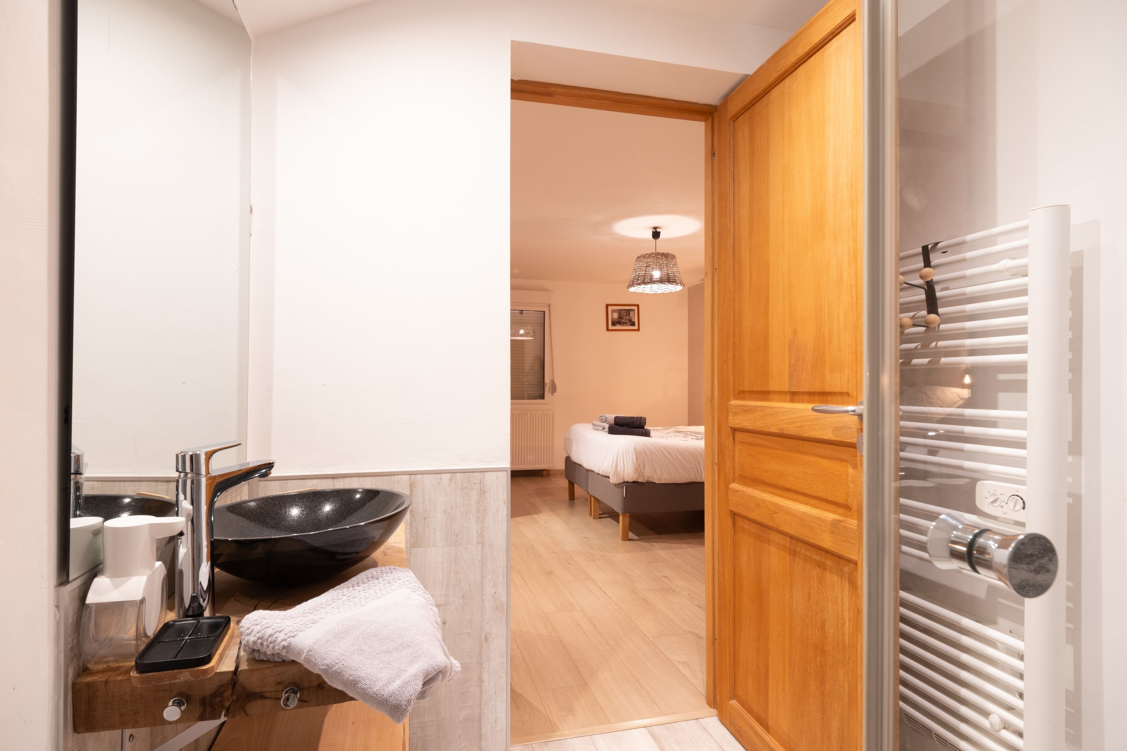A modern bathroom with a large mirror, a black basin, a towel, and a wooden door leading to a bedroom with a bed and a hanging light fixture.