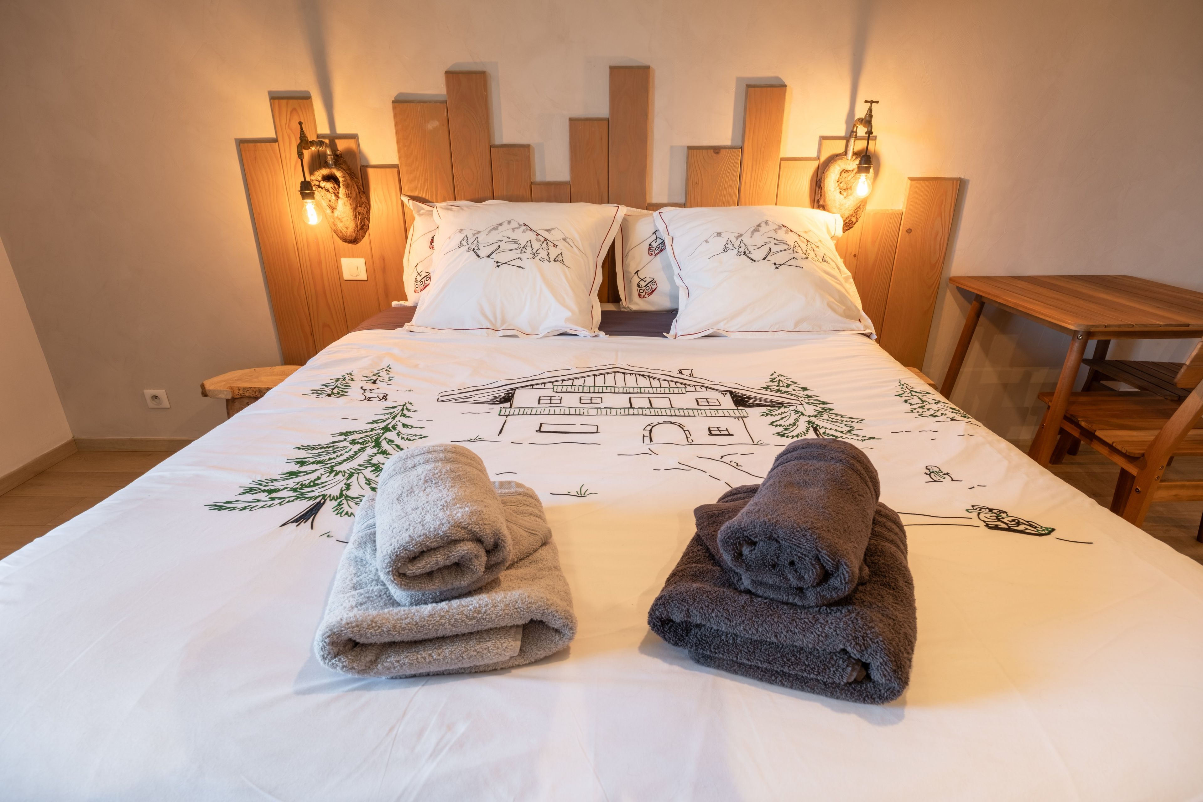 Cama bem arrumada com design de montanha e casa, duas toalhas e cabeceira de madeira com luzes de parede.