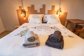 A neatly made bed with a mountain and house design, two towels, and wooden headboard with wall-mounted lights.