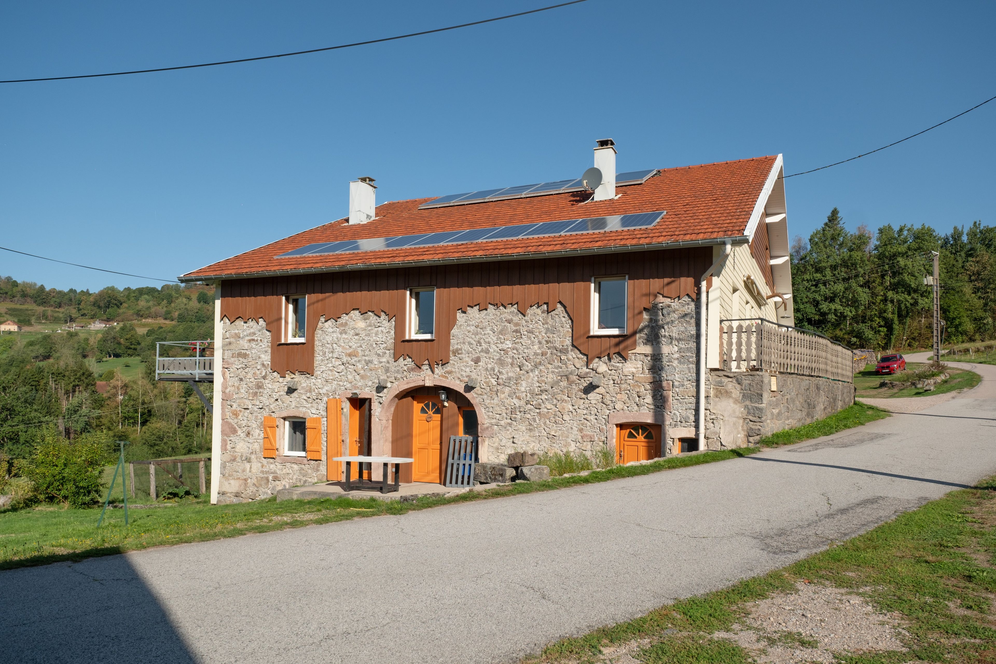 Een stenen huis met twee verdiepingen, voorzien van een rode dakbedekking, houten luiken en een klein tafeltje buiten.