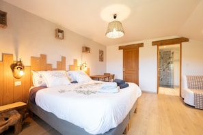 Un dormitorio con cama, mesitas de noche, cabecera de madera, lámpara colgante, puerta que da a un baño y una pequeña zona de estar.