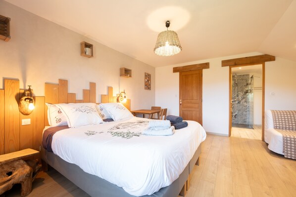 Un dormitorio con cama, mesitas de noche, cabecera de madera, lámpara colgante, puerta que da a un baño y una pequeña zona de estar.