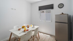 Una cocina pequeña y luminosa con una mesa blanca lista para el desayuno, un reloj en la pared y una ventana con persianas.