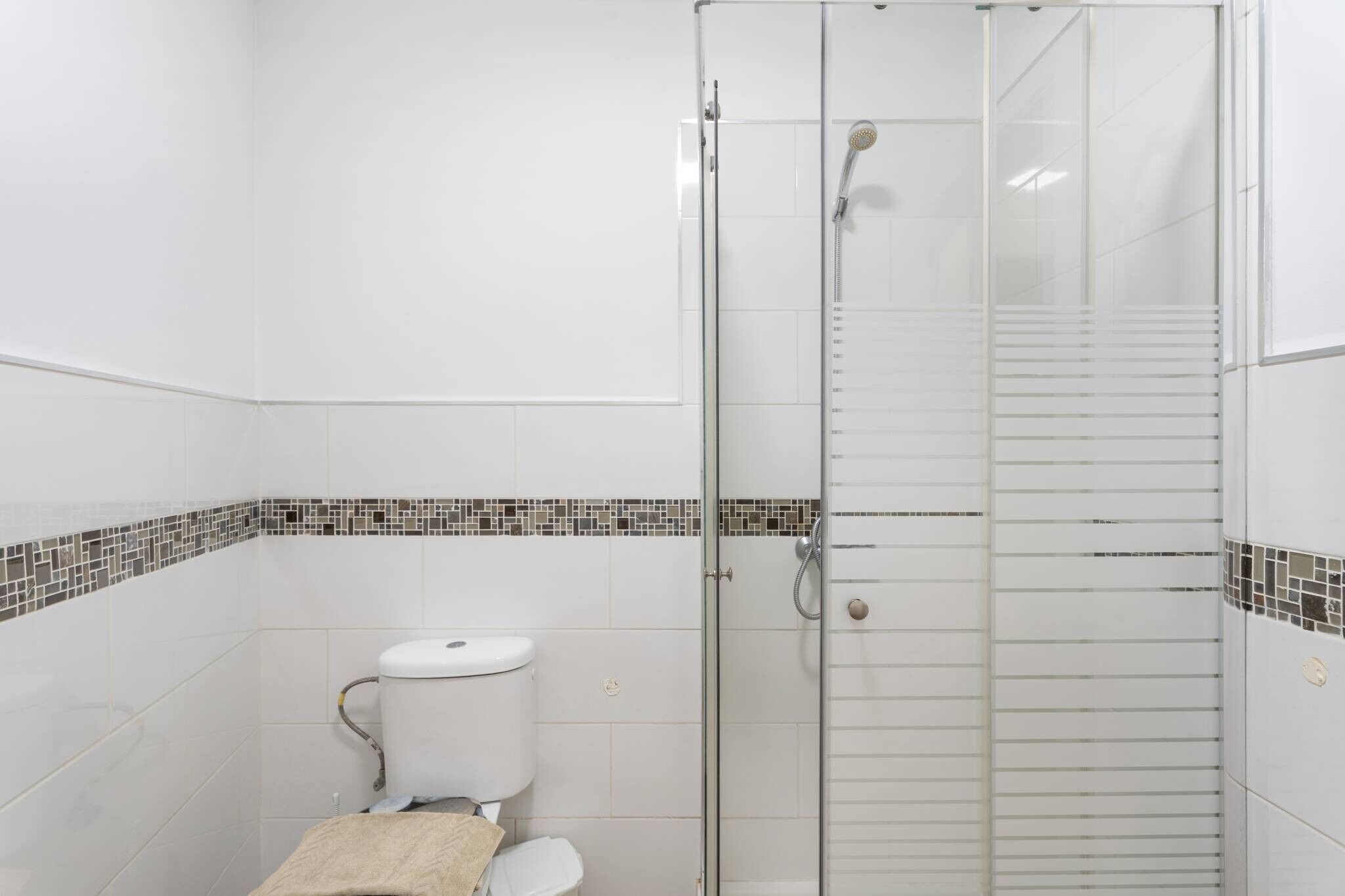 Een badkamer met een douchecabine van glas, een toilet en een betegelde vloer met decoratieve rand.