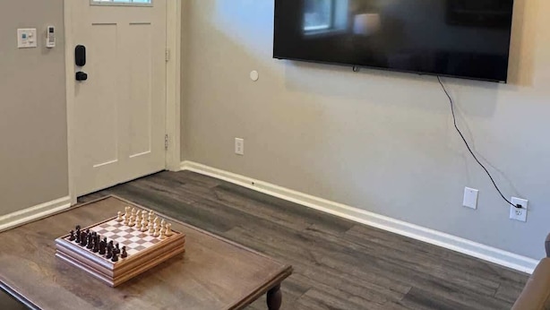 A room with a ceiling fan, a mounted flat-screen TV, a chessboard on a wooden table, and a door with a window.