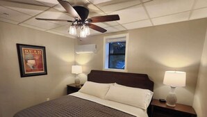 A bedroom with a bed, two bedside tables, a ceiling fan, a window, and a wall-mounted air conditioner.