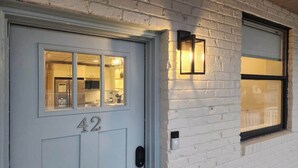 A white brick house with a number '42' on the door, a window with a black frame, and a wall-mounted light fixture.