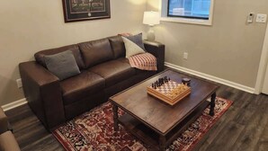 A living room with a brown leather sofa, a wooden coffee table with a chessboard, a patterned rug, a framed picture on the wall, and a window with a view.