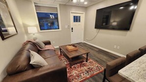 Una sala de estar con un sofá de cuero marrón, una mesa de centro de madera, un tapete estampado, un televisor montado en la pared y una puerta con ventanal.
