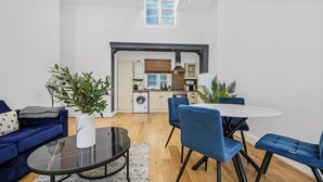 Ein modernes Wohnzimmer mit einem runden Glastisch, blauen Sofas und einem Essbereich mit weißem Tisch und blauen Stühlen. Im Hintergrund ist eine Küche mit integriertem Backofen und Mikrowelle erkennbar.