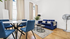 Ein modernes Wohnzimmer mit einem runden Esstisch, blauen Polsterstühlen, einem blauen Sofa mit gemusterten Kissen, einer Stehlampe und einem Standventilator.