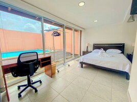 Habitación de hotel con cama, escritorio, silla, mesita, lámpara y vista a una piscina.