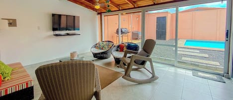 Een ruime woonkamer met een televisie, rieten meubels en uitzicht op het buitenzwembad.