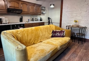 A yellow sofa in a room with a kitchen area, a dining table with chairs, and a white brick wall.