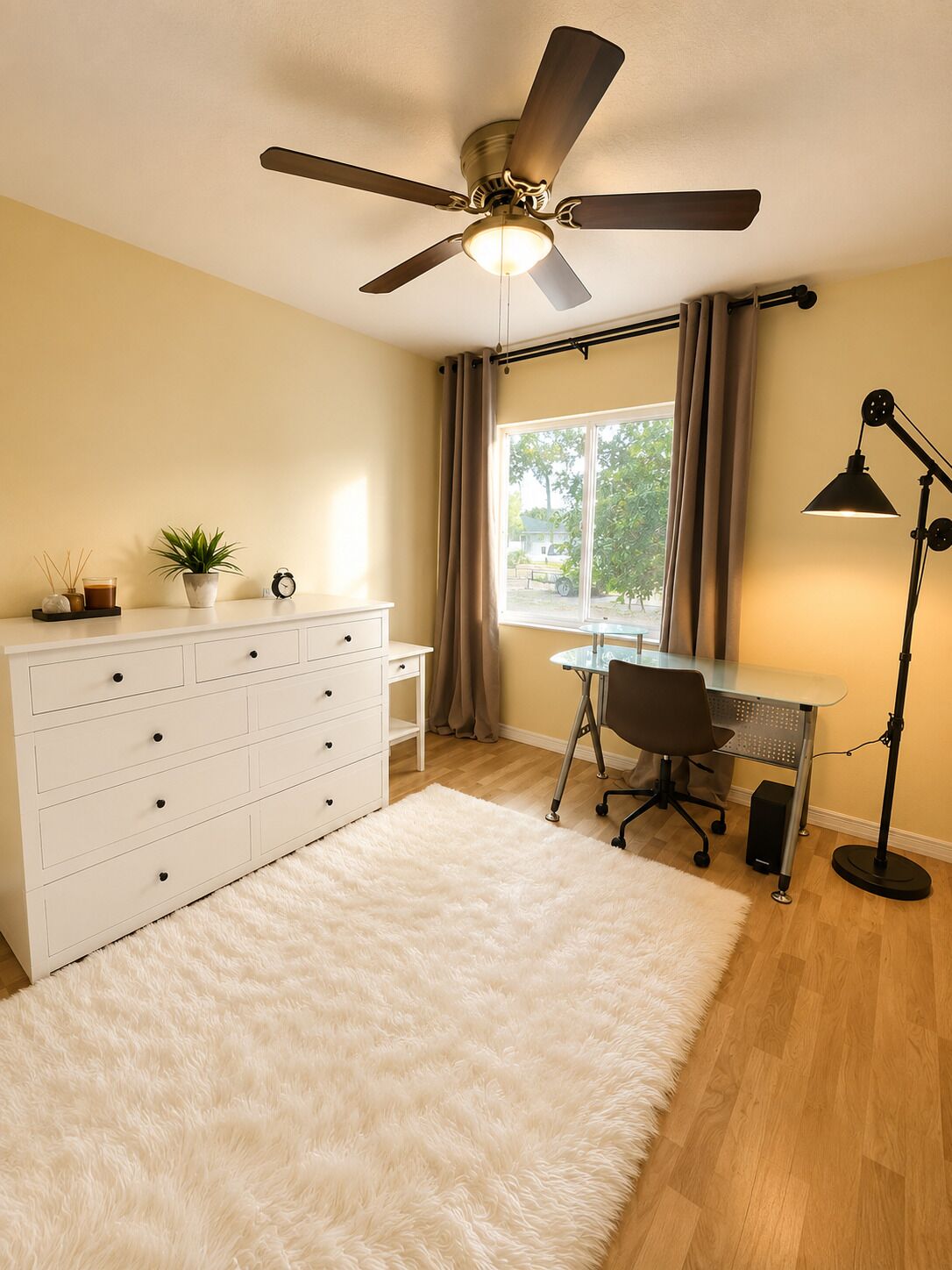A room with a ceiling fan, a desk with a chair, a white dresser, a window with curtains, and a white fluffy rug.