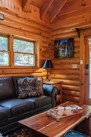 A cabin interior with a wooden ceiling, a black leather sofa with patterned cushions, a wooden table with cups and a lamp, and a painting of a waterfall on the wall. - Relax by the Waterfall ,Hot Tub,Pet-Friendly (Sevierville)