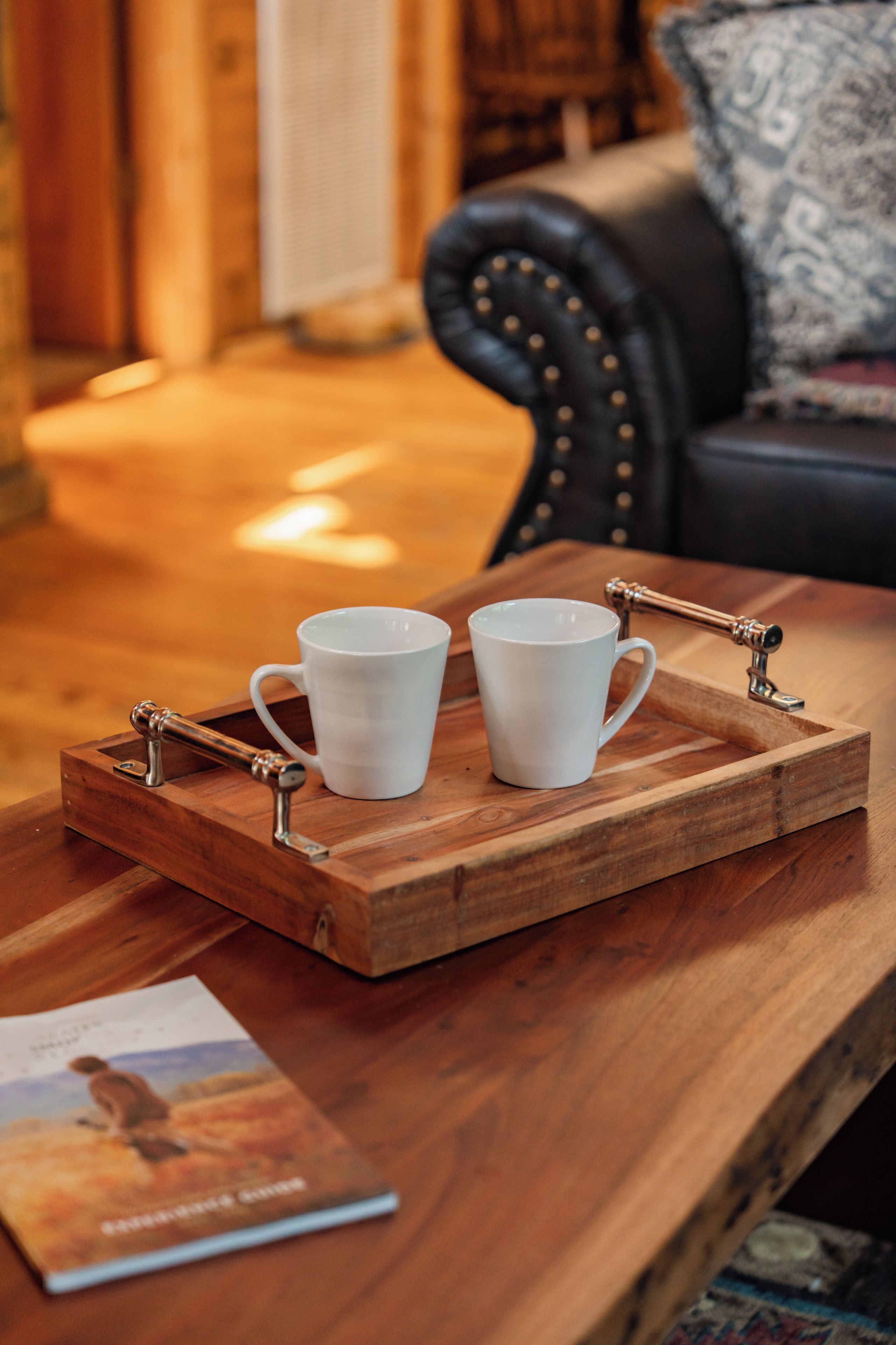 A wooden tray with two white cups on a wooden table, a book, and a patterned cushion in the background.
