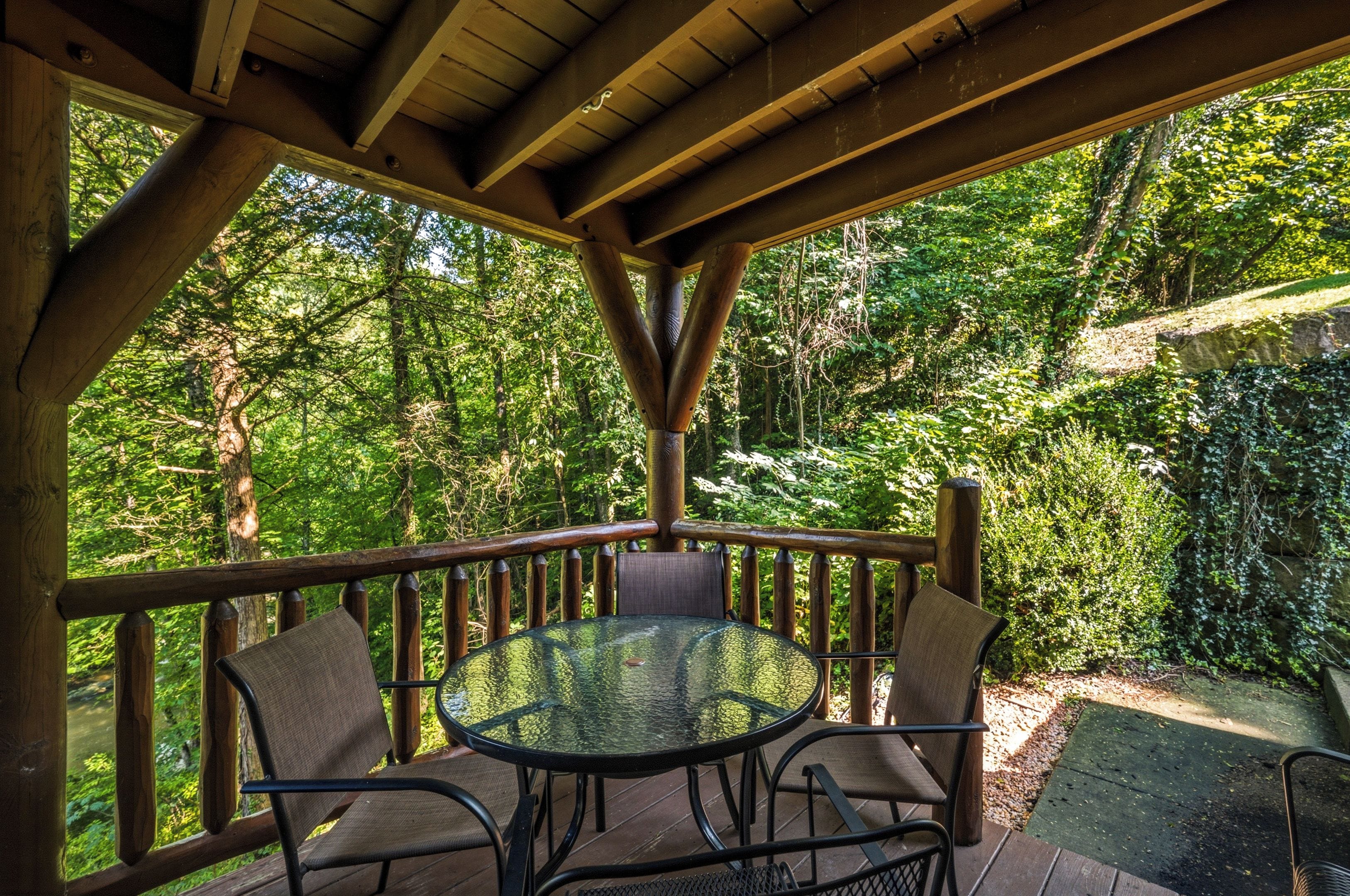 A covered outdoor seating area with a round glass table and four chairs, surrounded by a wooden deck and a view of a lush garden.