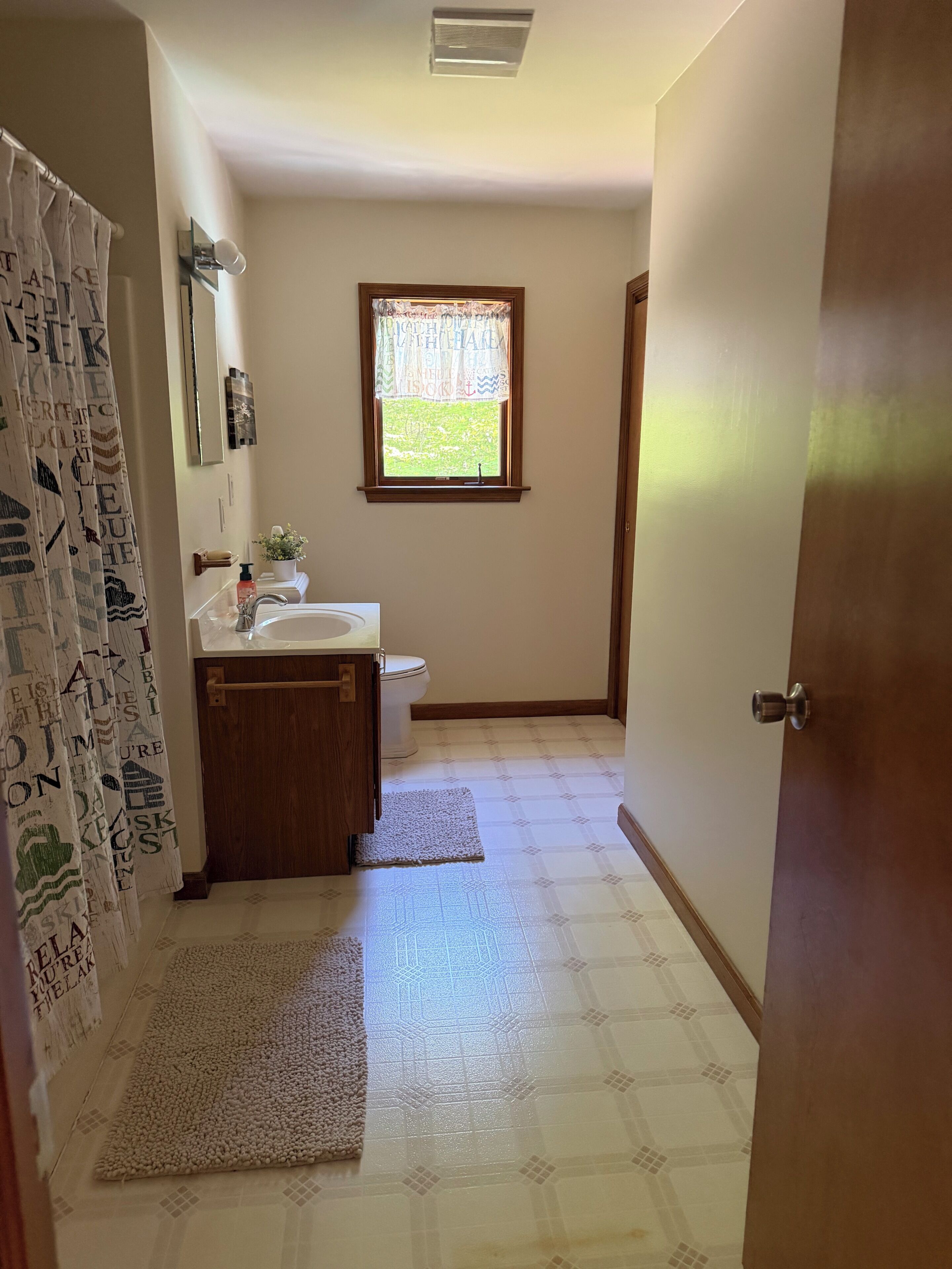 Ein Badezimmer mit Toilette, Waschbecken und Fenster. Zwei Teppiche liegen auf dem Boden.