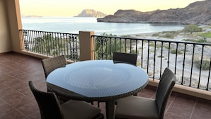 A balcony with a round glass table and wicker chairs, overlooking a coastal landscape with mountains and a body of water. - Luxury Two Bedroom Ocean Front Penthouse with Spa on Terrace at Villa del Palmar (Loreto)