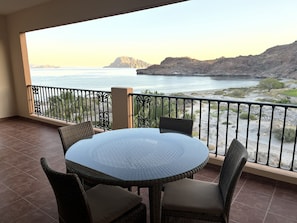 Een balkon met een ronde glazen tafel en rieten stoelen, met uitzicht op een kustlandschap met bergen en een wateroppervlak.