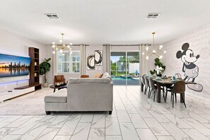 Una sala de estar moderna con un sofá gris, un comedor con mesa y sillas, un televisor grande y un mural de Mickey Mouse en la pared.