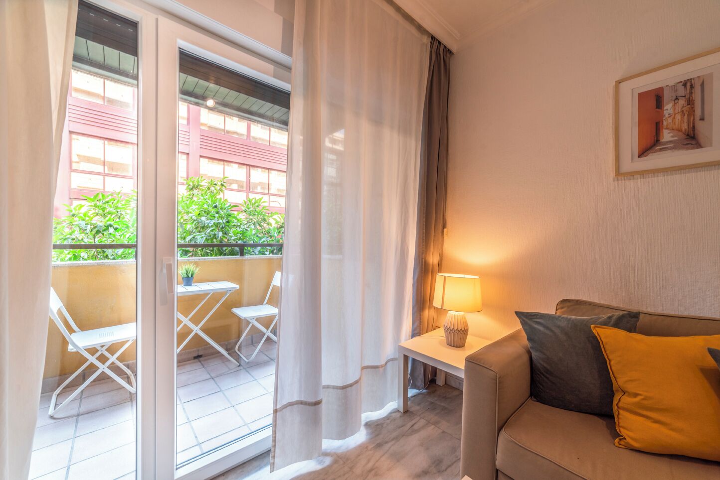 Ein Hotelzimmer mit Balkon, einer Couch mit Kissen, einem kleinen Tisch mit Lampe und einem gerahmten Bild an der Wand.