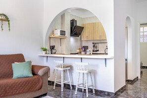 Ein gemütlicher Wohnraum mit Küchenzeile, einem braunen Sofa mit grüner Kissen und einem weißen Sessel.