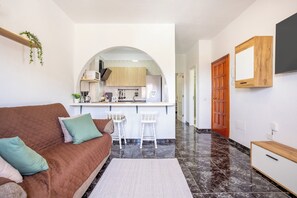 A living room with a brown sofa, a small kitchen area, and a hallway leading to a door. - Home2Book Ocean & Mountain View in San Andrés (San Andres de Tenerife)